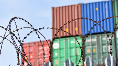 Opgestapelde zeecontainers achter een hek met prikkeldraad in een haven- of opslaggebied.